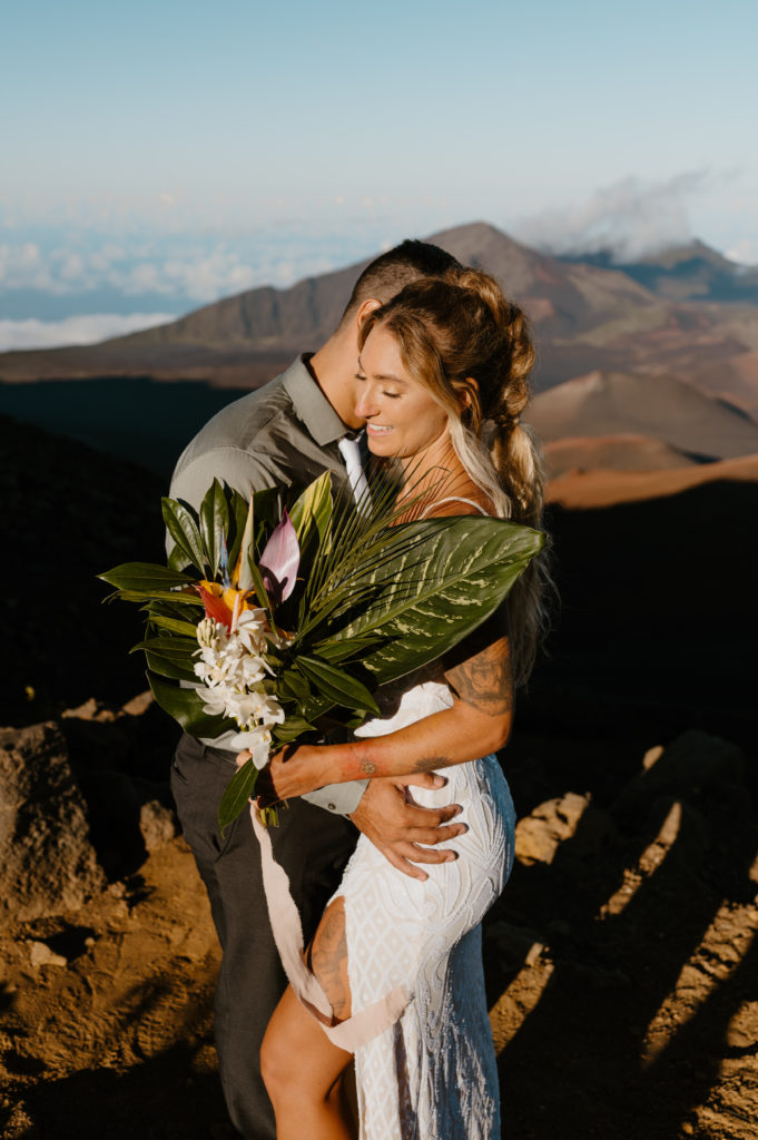 A couple embraces one another while holding a tropical floral arrangement in Maui