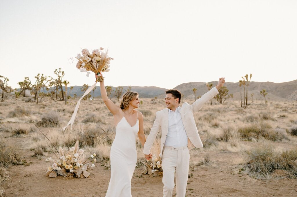 A couple in neutral wedding attire cheers in celebration of their elopement in Joshua Tree National Park