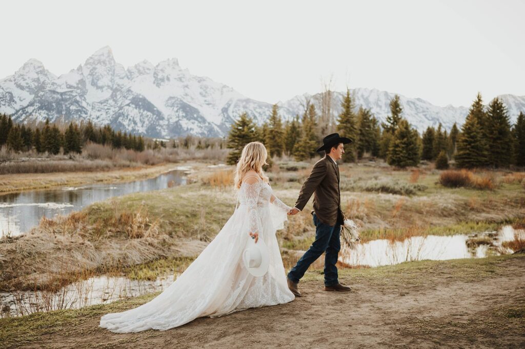 A husband leads his wife along a water's edge in the mountains after their wedding