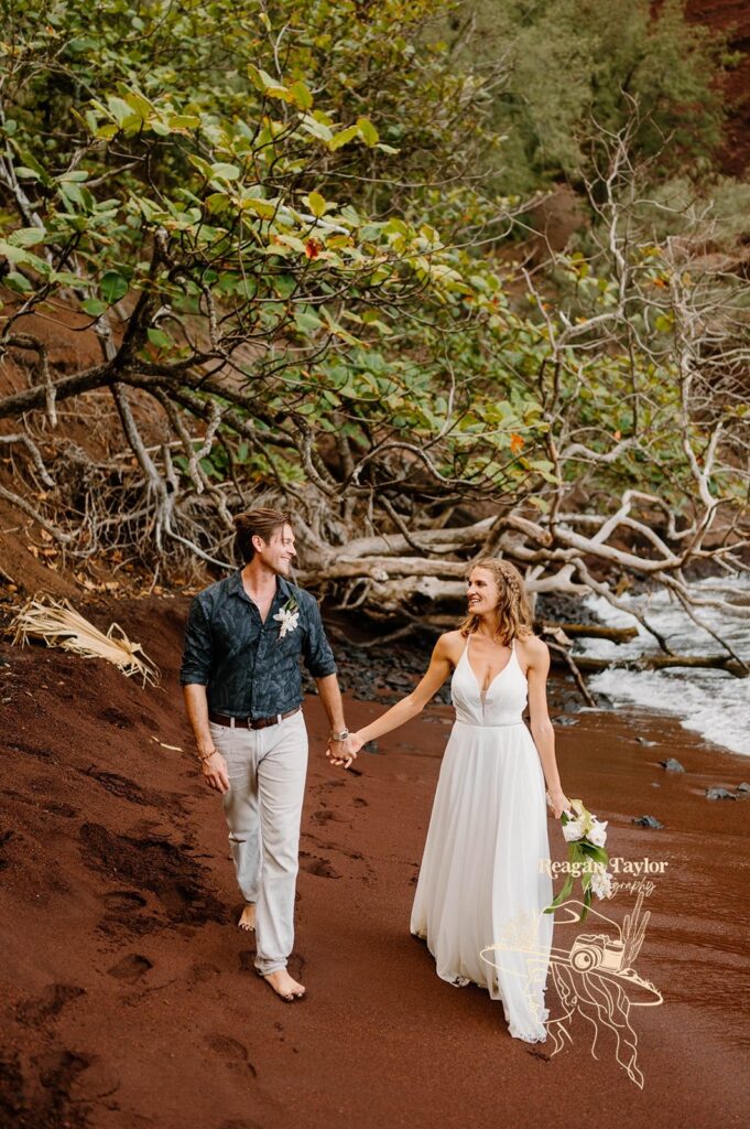 A couple holds hands while walking on red sand beach with wedding florals