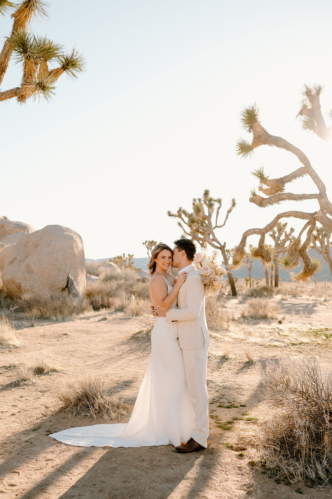 A woman smiles while her husband kisses her forehead during their Joshua Tree elopement at Sunset
