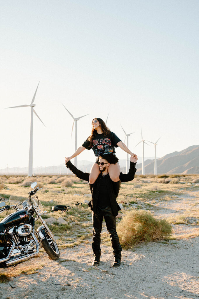 A woman playfully sits on her husband's shoulders as they explore and adventure a field of windmills near Palm Springs