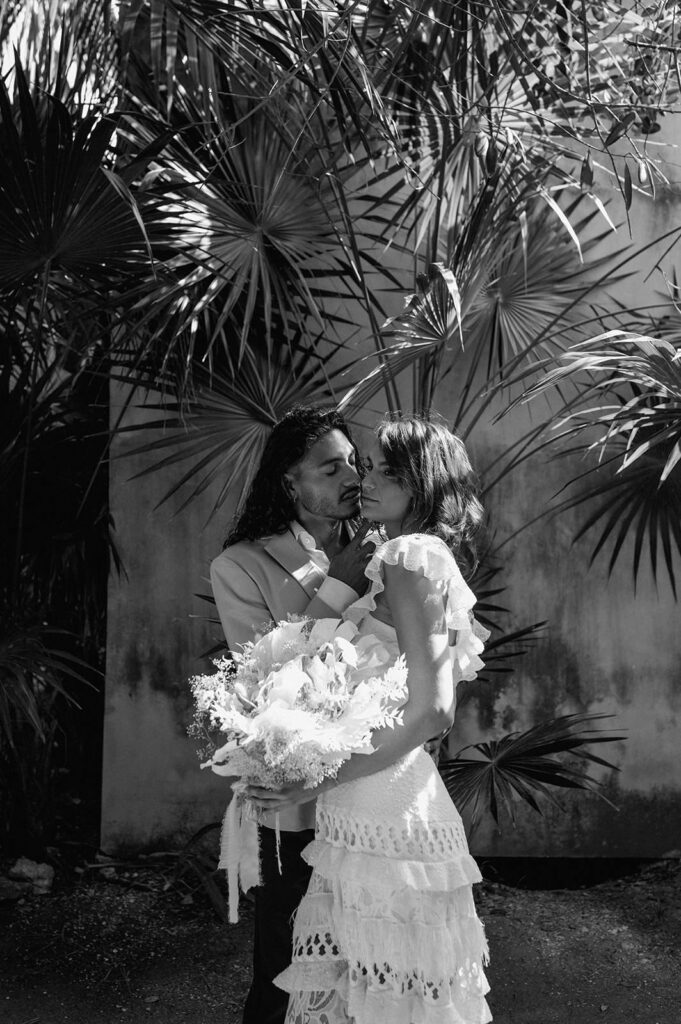 A newlywed couple kisses while holding wedding florals in Tulum near a palm tree