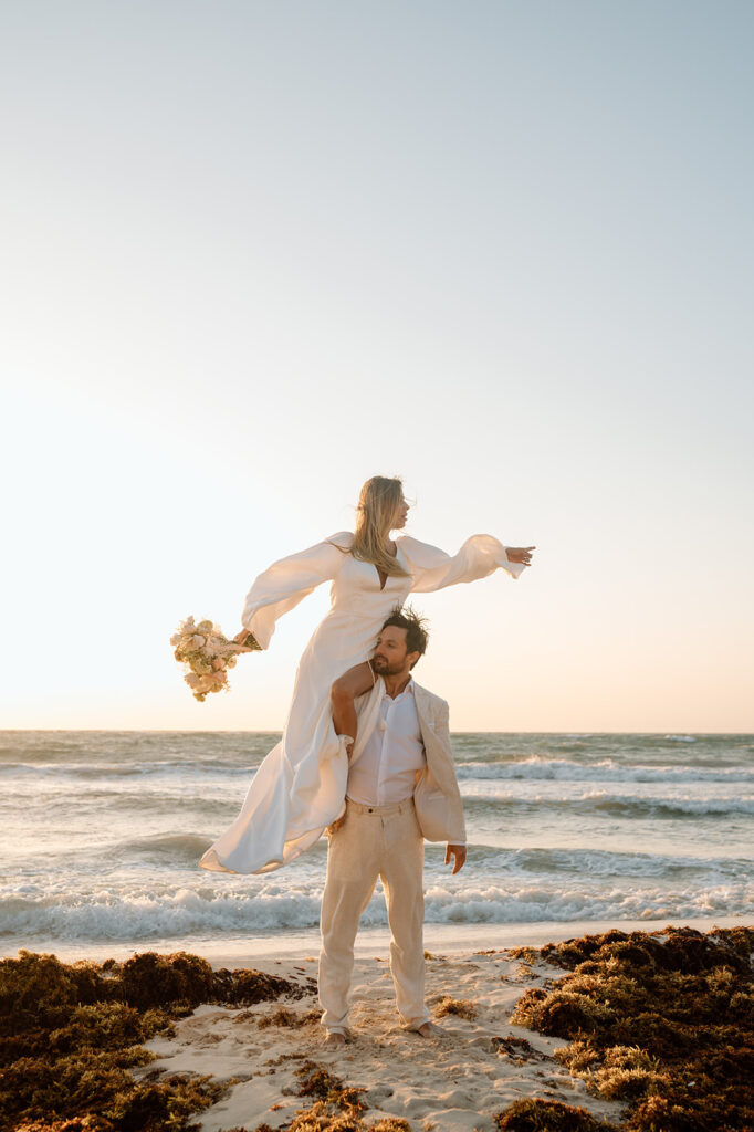A woman is on a man's shoulder holding wedding florals on a sandy beach elopement