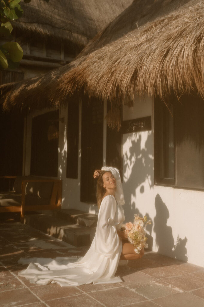 A woman poses on thee ground with her hand in her hair in a wedding dress at a cabana in Tulum