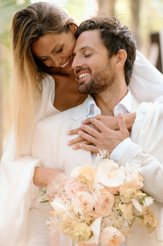 A bride hugs her husband while he is sitting with wedding florals in hand