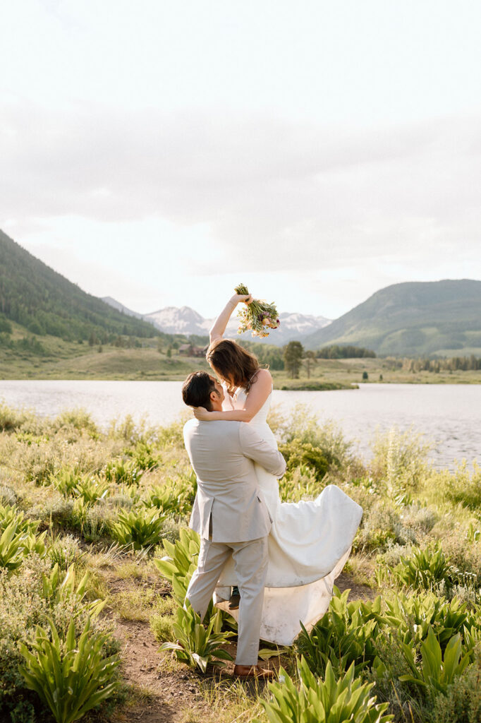 A bride is picked up by her groom with wedding florals in hand near an alpine lake.