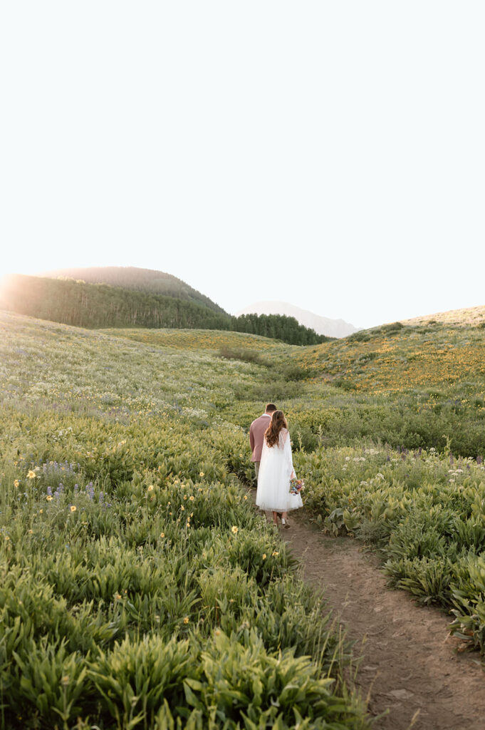 A bride and groom stroll along a dirt path while passing wildflowers