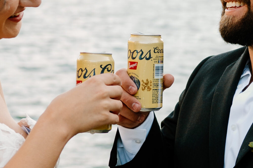 A newlywed couple shares a Coors Banquet by an alpine lake. 