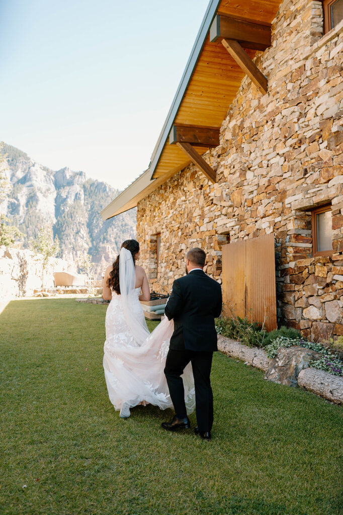 A bride's groom helps her carry her dress train up a grassy hill at a mountain lodge.