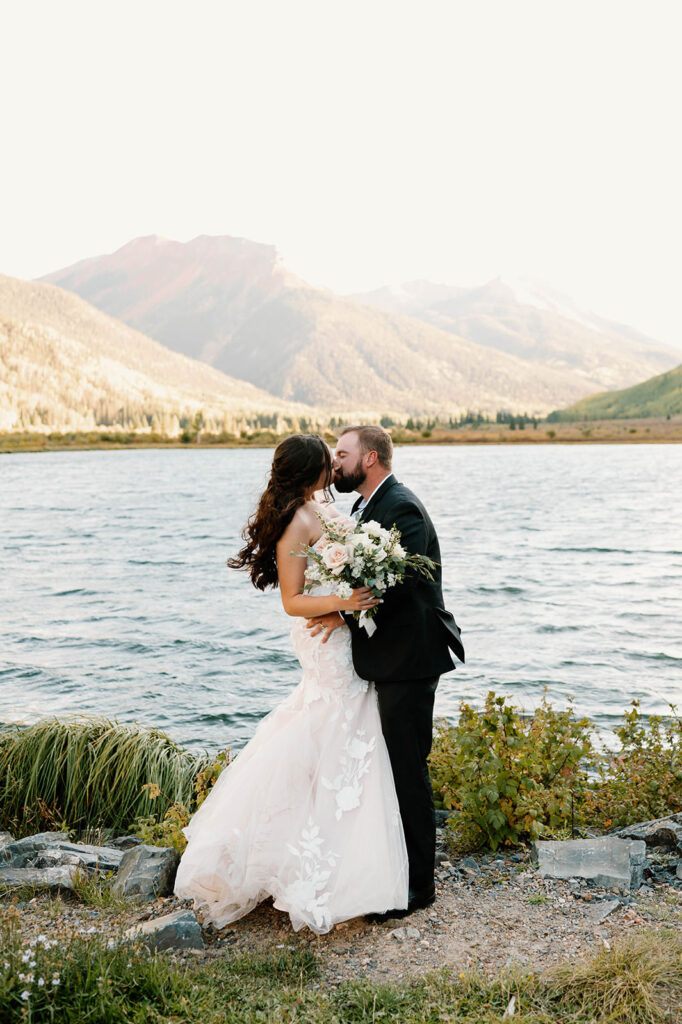 A newlywed couple kisses by an alpine lake near the San Juan Mountains. 