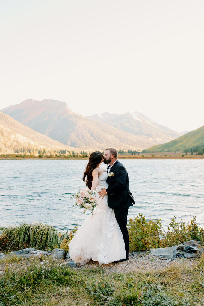 A couple celebrates their recent marriage with a kiss near an mountain lake. 