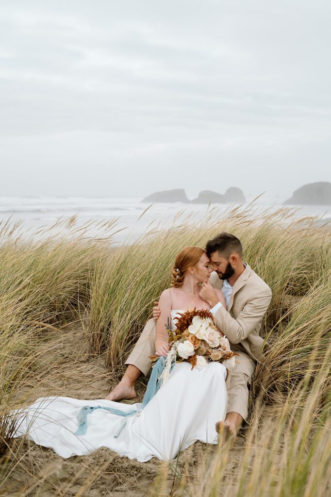 A couple sits in tall grass near the sea during their wedding portraits.