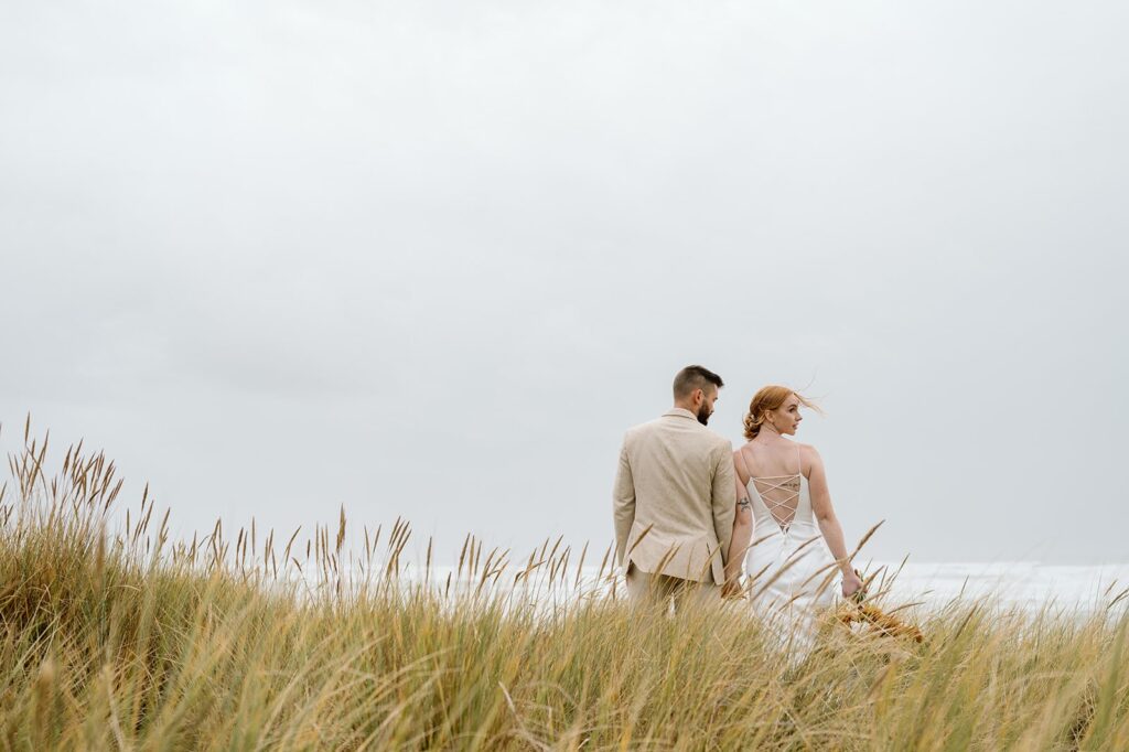 A couple walks through sand along Cannon Beach while holding wedding florals. 