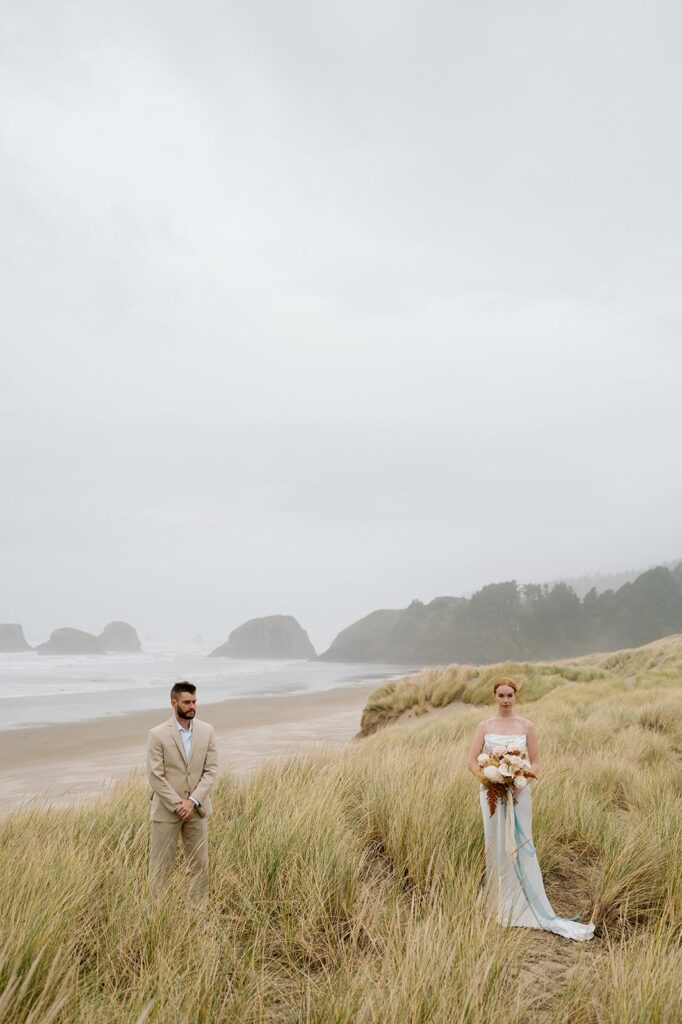 A newlywed couple stands in a meadow of seagrass on Cannon Beach in Oregon.