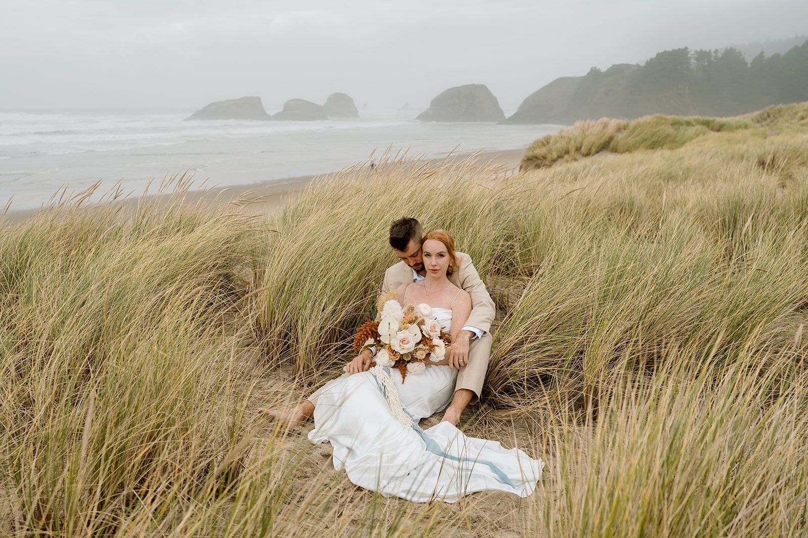 A couple sits in tall sea grass on the Oregon Coast during their Cannon Beach elopement.