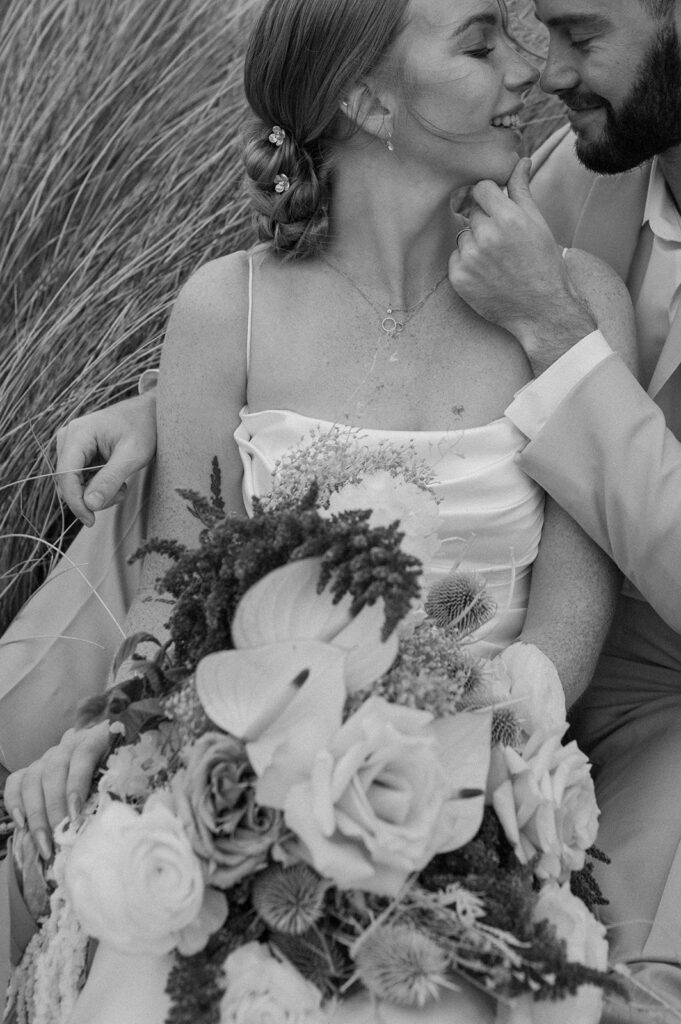 A couple relaxes in seagrass while holding neutral wedding florals. 