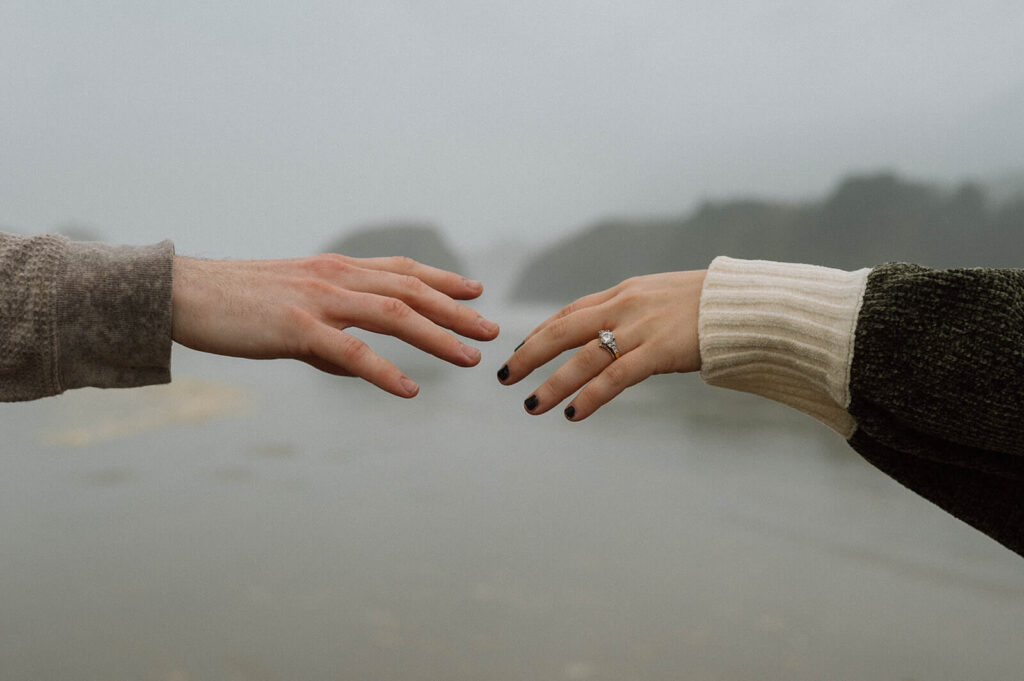 A couple's hands are stretched out to one another on a beach in Oregon.