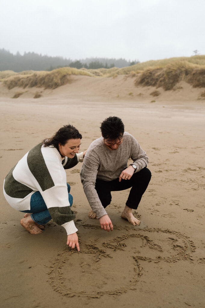 A couple draws in the sand during their beach engagement shoot. 