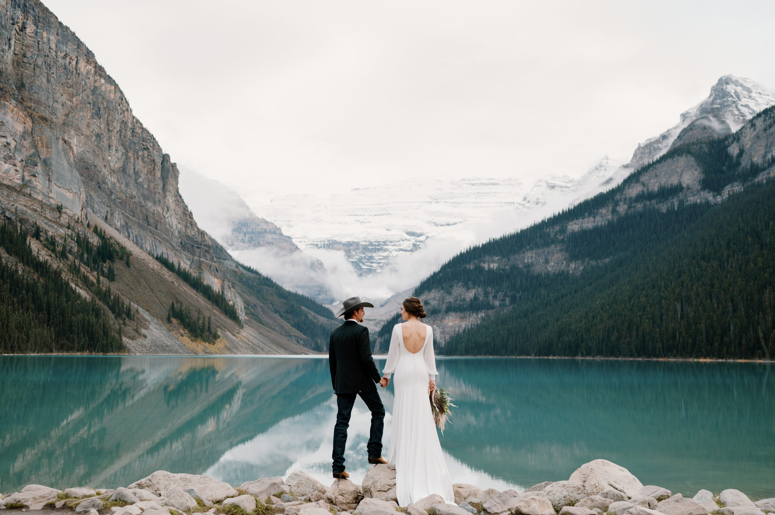 A couple stands on a pile of rocks that overlooks Lake Louise during their intimate Banff elopement.
