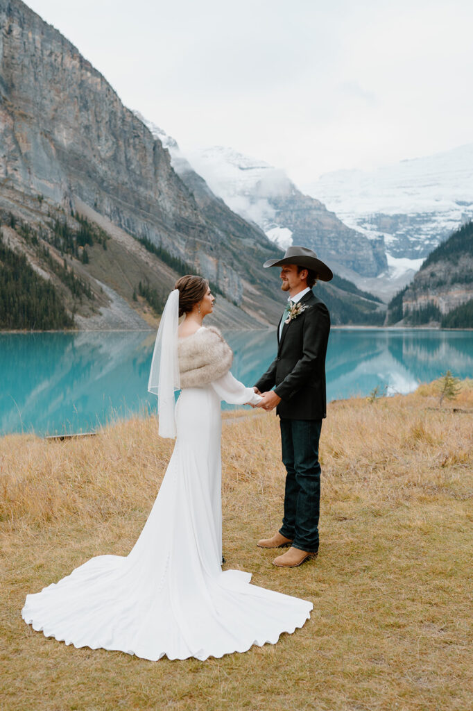 A woman in a long wedding gown and fur bridal wrap holds her groom's hands, who is wearing cowboy boots, a cowboy hat and a suit coat near Lake Louise. 