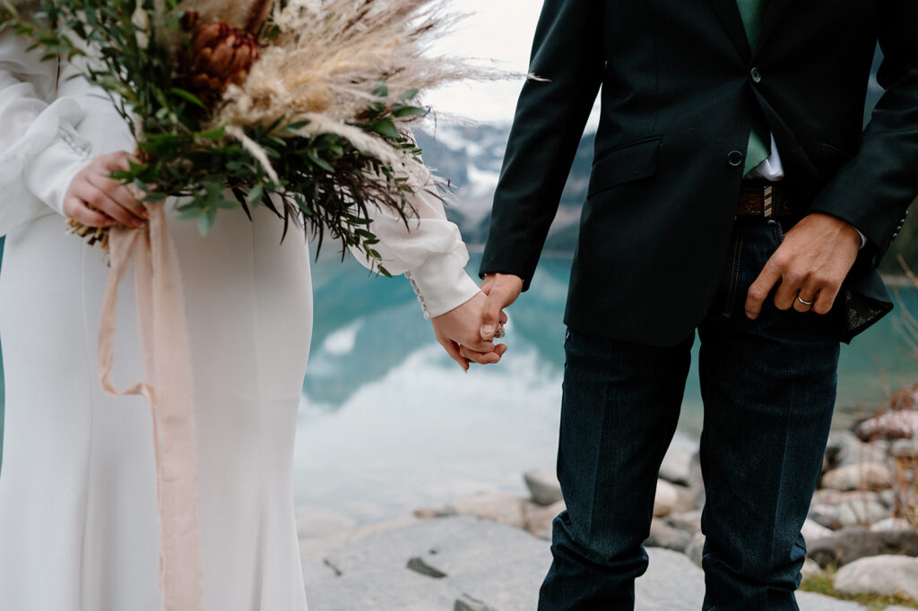 A bride, holding a bridal bouquet from Adventure Floral, grabs her groom's hand while they stand near Lake Louise. 