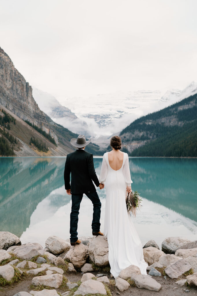 An adventure elopement couple holds hands while admiring a blue alpine lake in Banff National Park. 