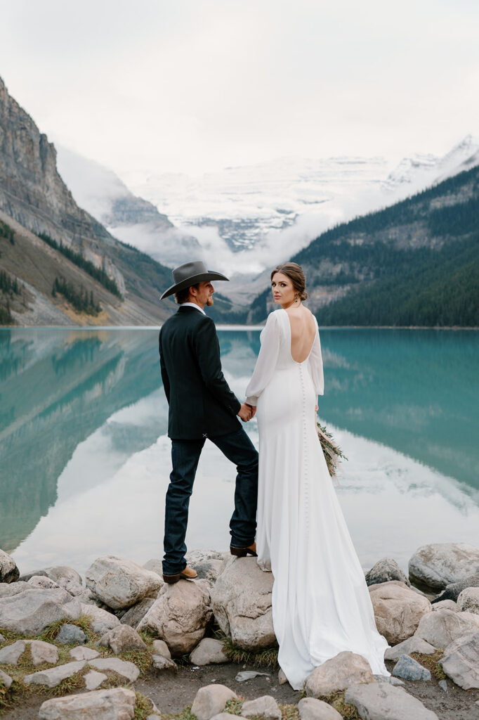 A Banff elopement couple stands on a pile of rocks along the shores of Lake Louise with snow capped mountains in the distance 