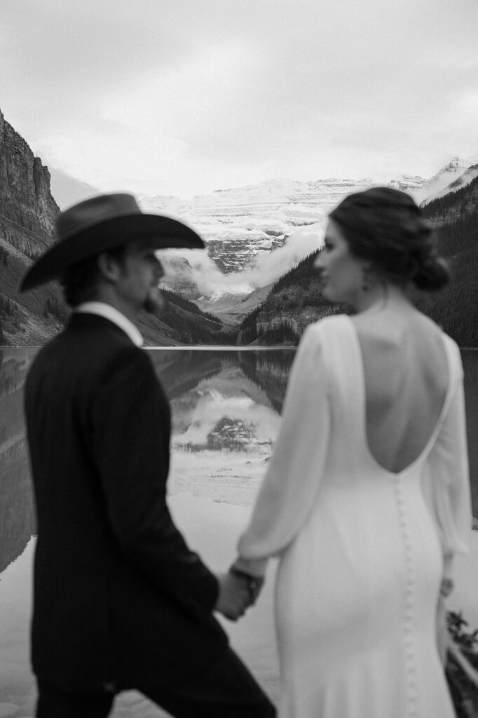 A bride, wearing a long sleeved wedding gown, looks at her groom, wearing a cowboy hat, with Lake Louise in the background. 