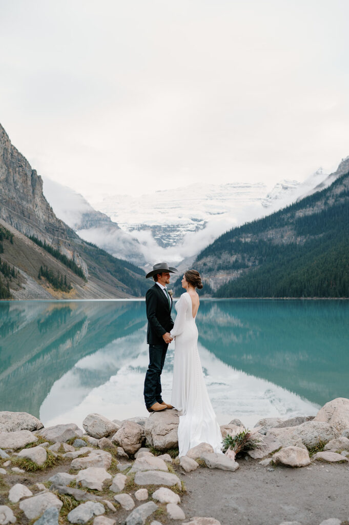A couple holds hands with one another during their Lake Louise elopement while standing on a bed of rocks along the turquoise lake's shoreline. 