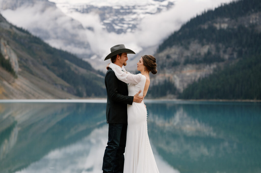 An man in a suit coat, jeans and cowboy hat stands with a woman in a wedding dress on the shores of Lake Louise during their Banff elopement. 
