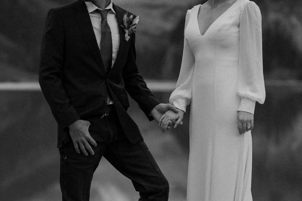 A man in a suit coat and jeans, and a woman in a long sleeve wedding dress, hold hands in Banff National Park. 