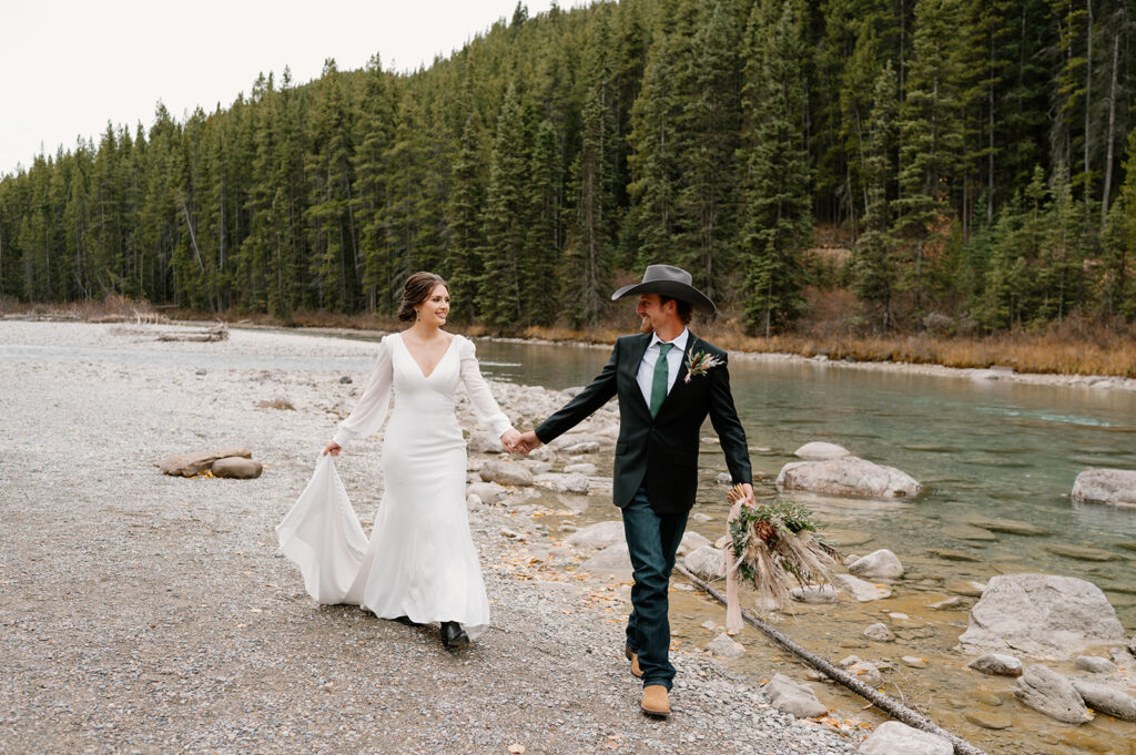 A bride is holding her groom's hand while walking along a stream in Banff National Park near Lake Louise. 