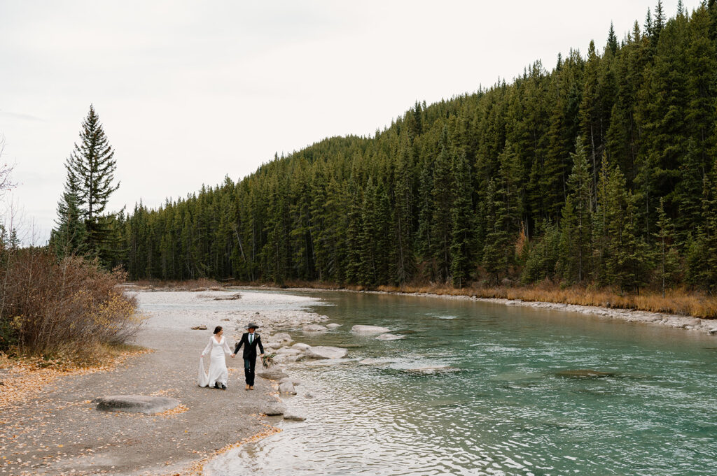 A bride and groom walk along a stream while holding hands in Banff National Park near Lake Louise. 