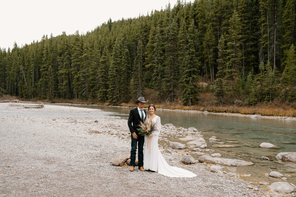 An adventure elopement couple stands for an outdoor wedding portrait near a stream by Lake Louise in Banff. 