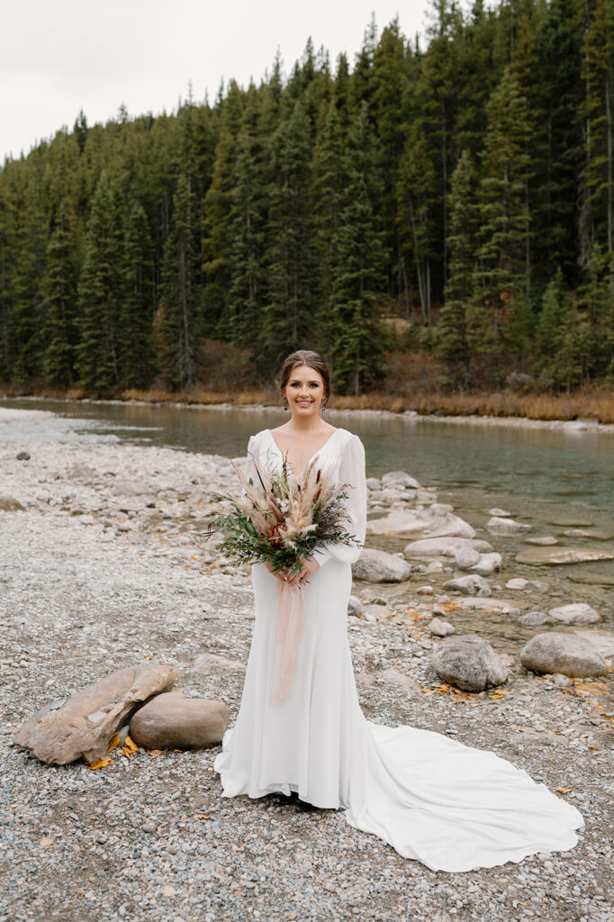 A bride, holding a floral bouquet from Adventure Floral, is wearing a long sleeved, white, floor length wedding gown near a stream in Banff.