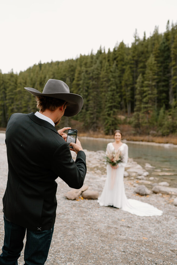 A groom takes a photo with an iPhone of his bride near a stream at Lake Louise for their elopement. 