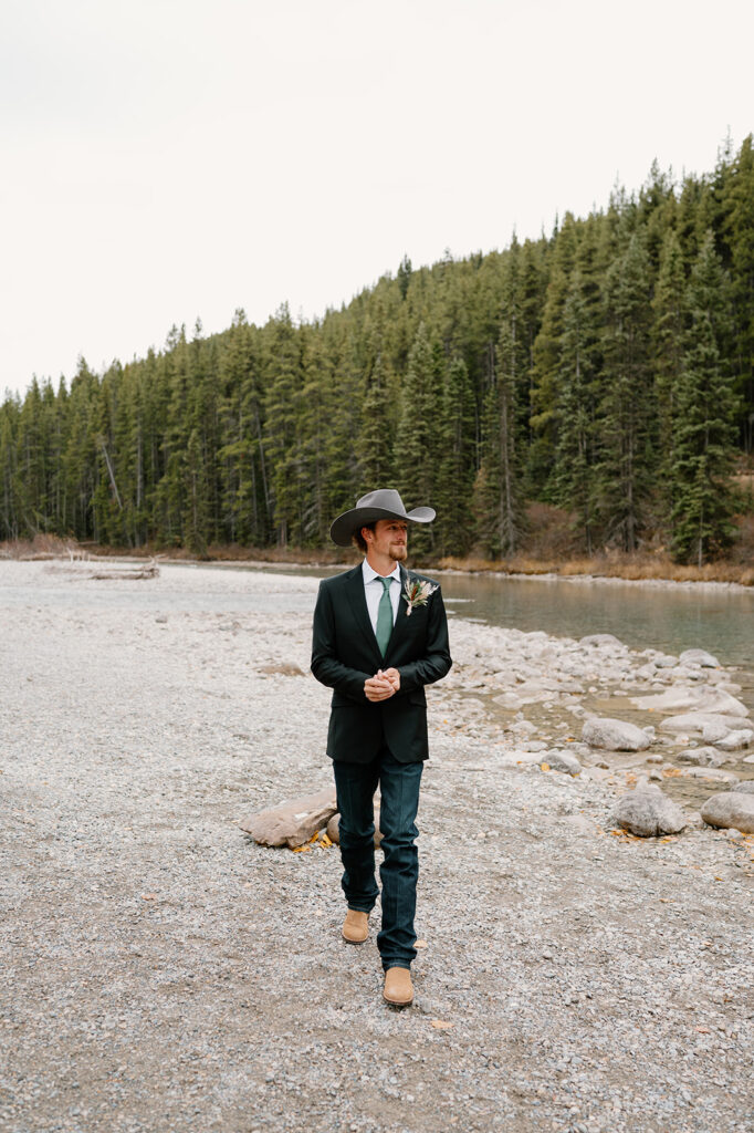 A groom, wearing a suit coat, dark jeans, light tan cowboy boots and a grey cowboy hat poses for a portrait near a stream in Banff.