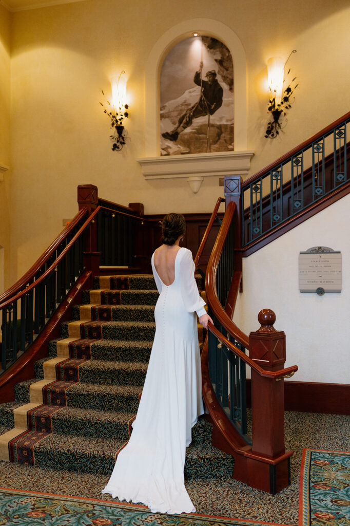 A bride in a long white wedding gown stands on a staircase with her back towards the camera at the Fairmont Chateau Lake Louise.