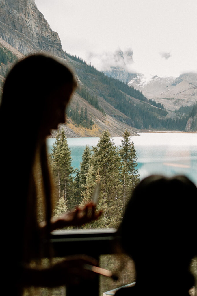 A bride sits in a Banff hotel that looks out onto an alpine lake while getting ready for her Banff elopement.