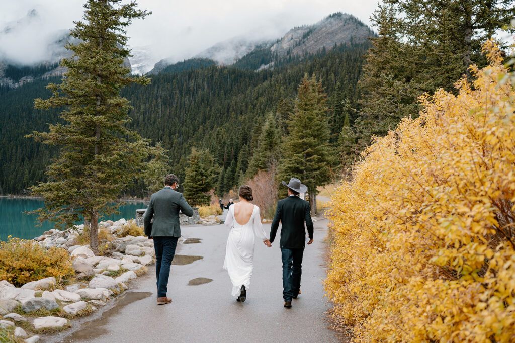 A couple and their officiant, Cole, from Married By Cole, walk along a path next to Lake Louise before celebrating a Banff elopement. 