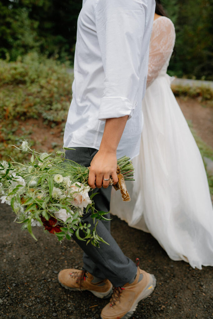 A groom holds his bride's floral bouqet while hiking in Oergo