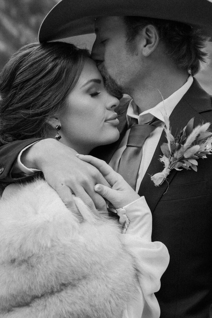 A groom, wearing a suit coat with a boutonniere on the lapel, kisses his bride's forehead, who is wearing a fur bridal wrap and drip earrings. 