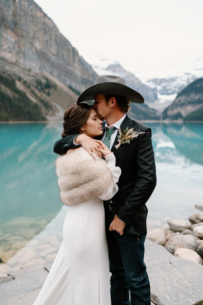 A woman in a fur shawl is held by a man in a suit coat and cowboy hat at Lake Louise during their Banff elopement. 