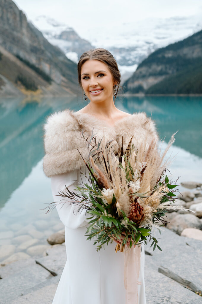 A bride with an updo, wearing a long sleeve white wedding gown holds a dried floral bouquet from Adventure Florals at Lake Louise. 