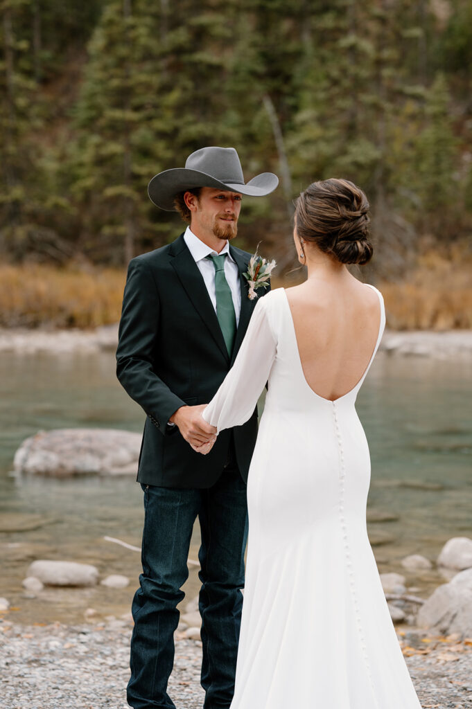 A groom in a grey cowboy hat, jeans and a suit coat holds his bride's hands during their first look by a stream in Banff.