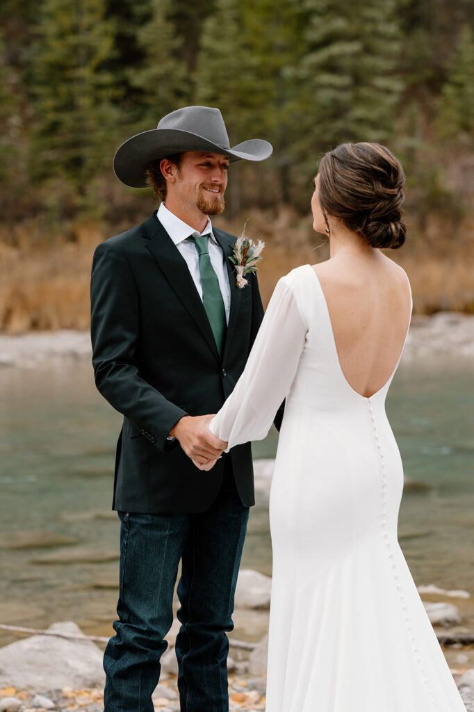 A bride in a long white wedding gown holds hands with her groom during their first look by a stream in Banff.