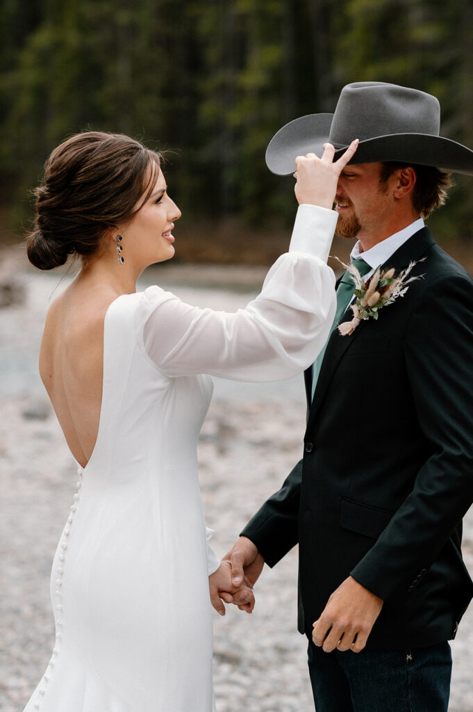 A bride in a white, long sleeve wedding gown adjusts her groom's grey cowboy hat before their Banff elopement. 