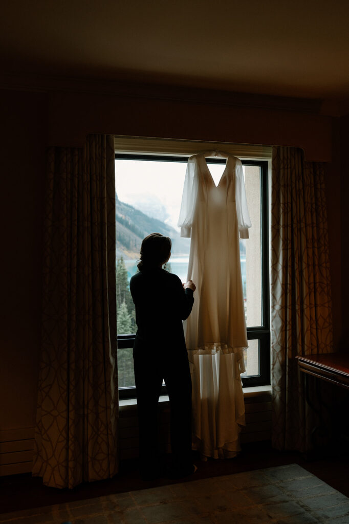 A bride looks at her wedding gown that hands from a hotel window looking out onto Lake Louise.