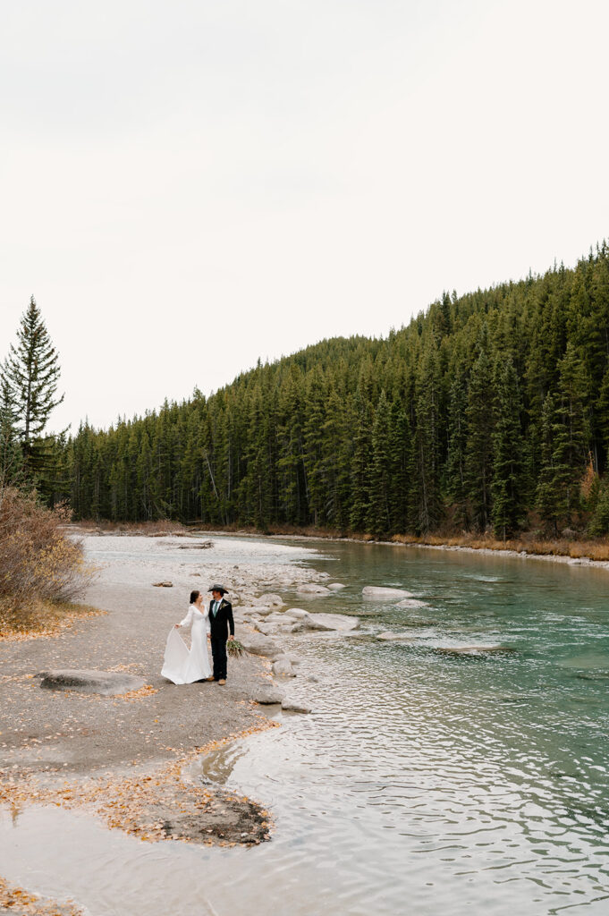 A woman wearing a long white wedding dress and a man wearing a suit coat, cowboy boots and a cowboy hat stand near an alpine lake stream in Banff. 